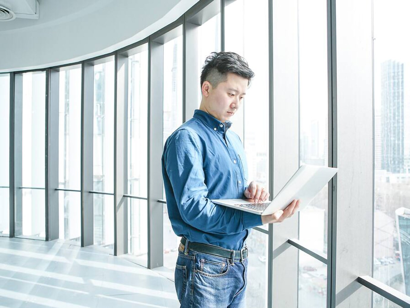 person looking at the laptop while standing near a window of a skyscraper